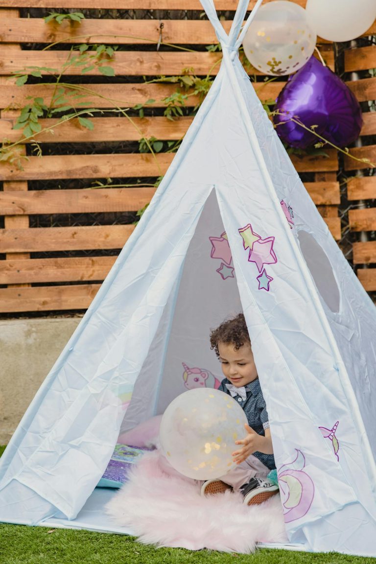Adorable child sitting inside a decorative teepee tent with balloons outdoors in a yard.